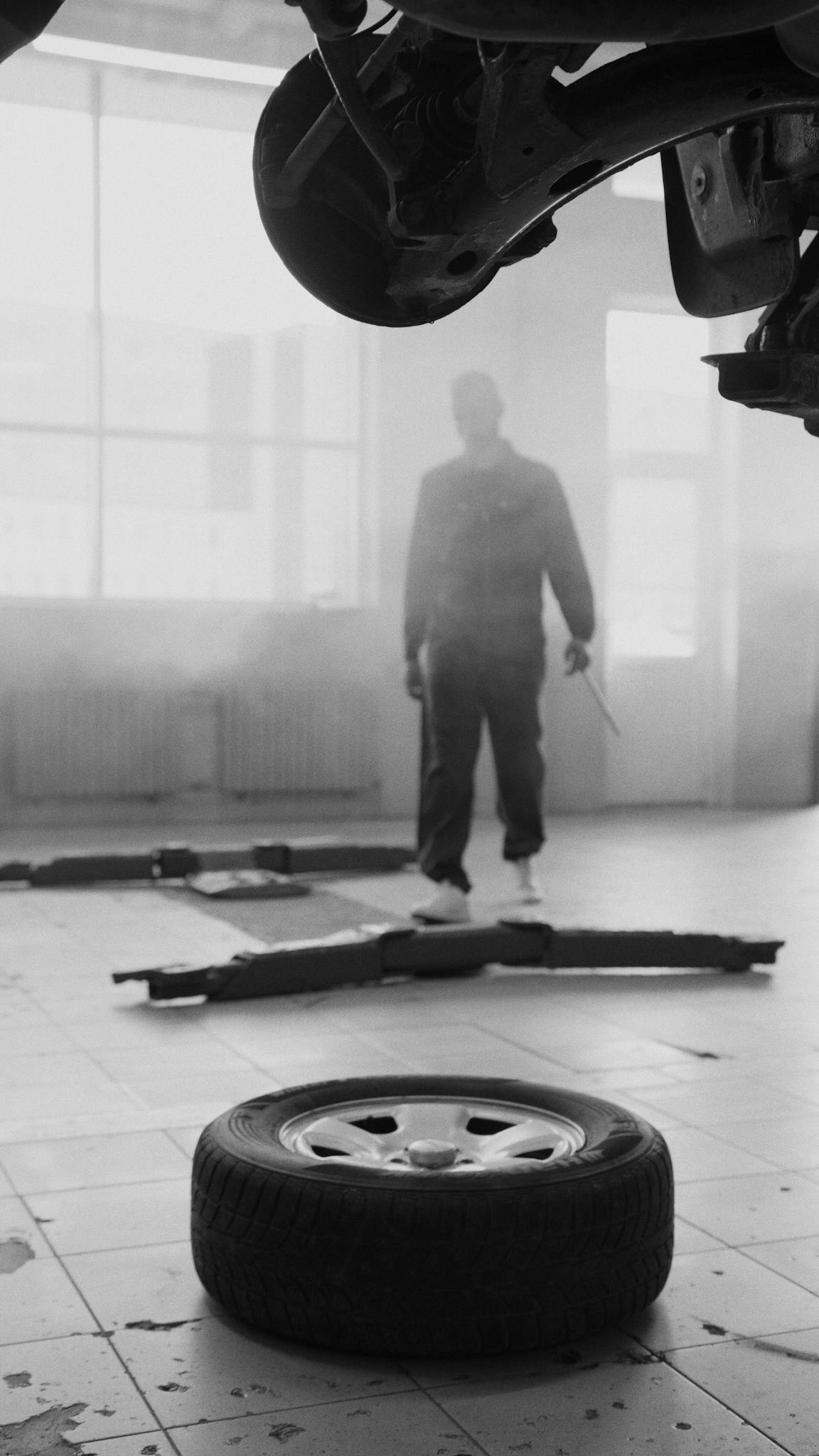 Black and white workshop scene with a mechanic examining a car. Focus on tire and equipment.